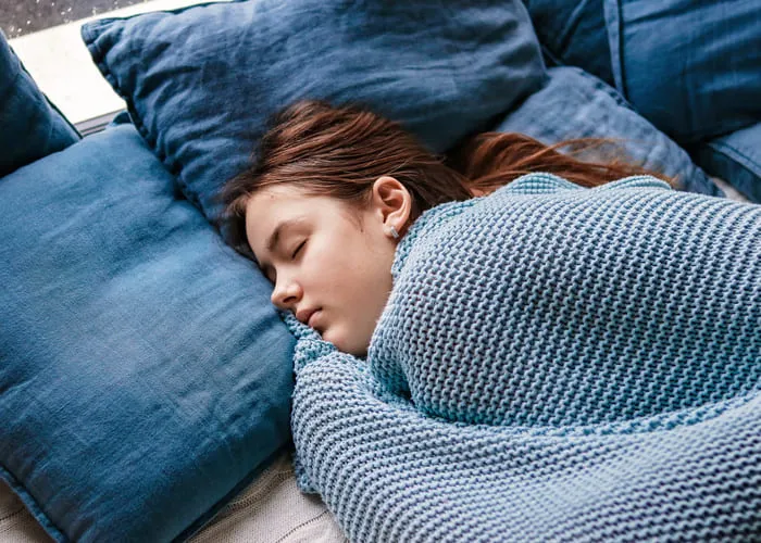 Young teenager girl sleeping snuggled in warm knitted blue blanket. Seasonal melancholy, apathy and winter blues. Cozy home.